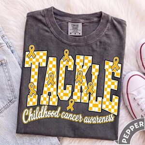 May include: A charcoal gray t-shirt featuring the word TACKLE in a yellow and white checkered pattern with yellow awareness ribbons integrated into the letters. Below, the text Childhood cancer awareness is printed in a white script font.
