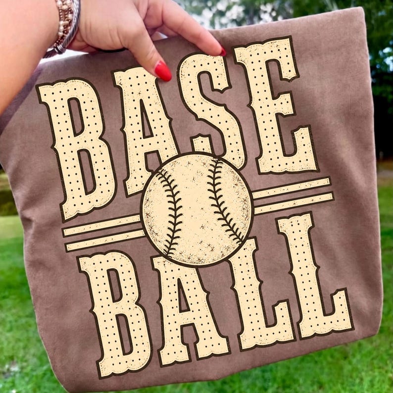 Puede incluir: Una camiseta marr&oacute;n con un gran estampado gr&aacute;fico que presenta la palabra BASE en una fuente de color crema con puntos sobre una ilustraci&oacute;n central de b&eacute;isbol, seguida de la palabra BALL en el mismo estilo de fuente, todo con l&iacute;neas.