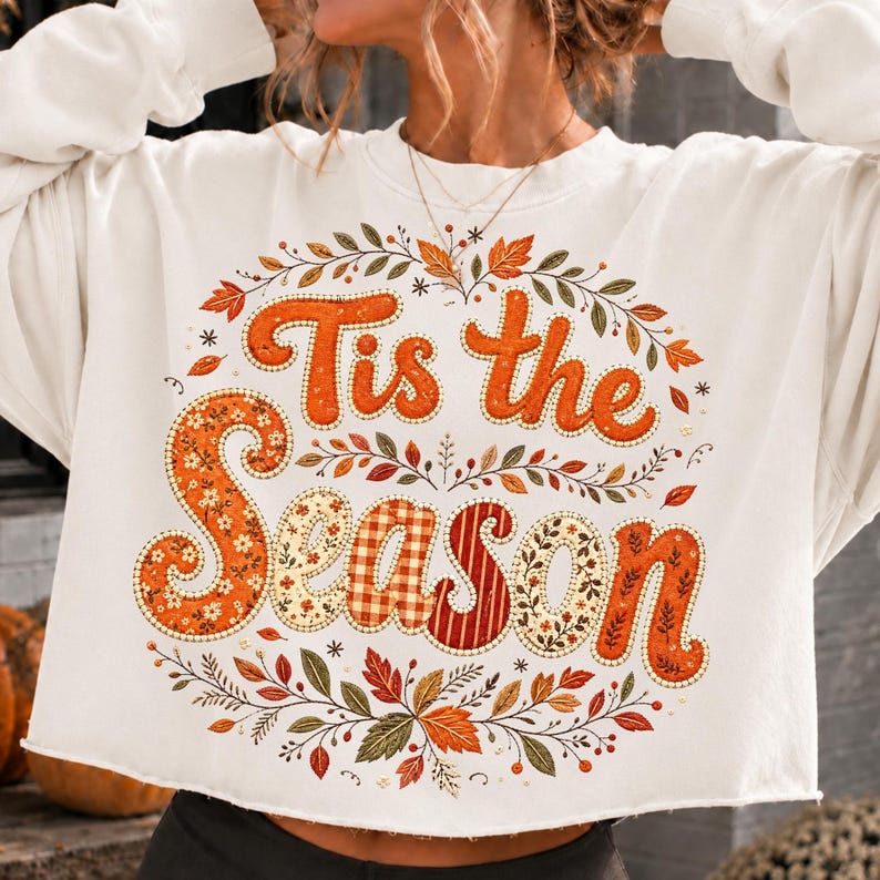 May include: A white long-sleeve sweatshirt featuring the text "Tis the Season" in large orange letters. The letters are filled with various patterns including floral, gingham, and stripes. The text is surrounded by a wreath of autumn leaves.