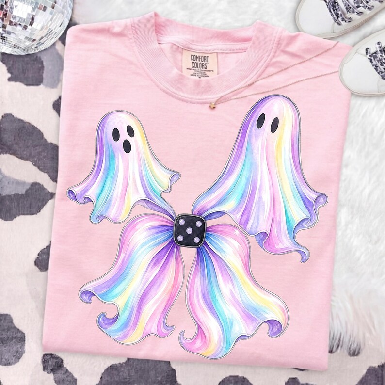May include: A light pink t-shirt featuring a graphic of two iridescent, pastel-coloured ghosts with black eyes and mouths, arranged to resemble a bow shape with a black die at the centre. The shirt has a Comfort Colors brand label at the neck.