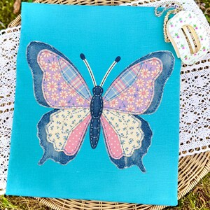 May include: A turquoise fabric square features a large butterfly applique. The butterfly has denim blue edges, pink and lavender wings with floral and plaid patterns, and a dark blue body. A small, floral-patterned item with a chain is in the upper right corner.