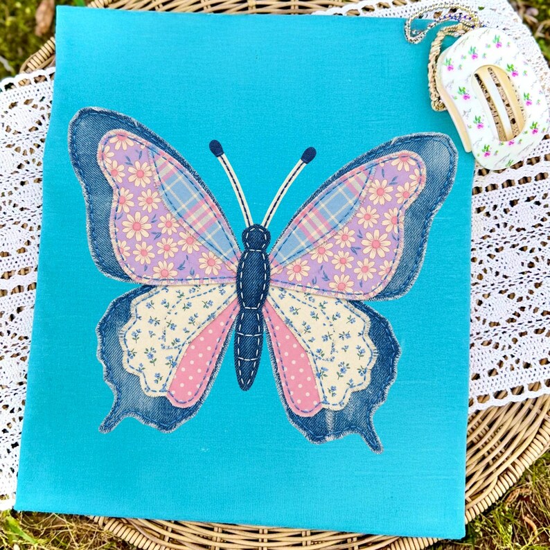 May include: A turquoise fabric square features a large butterfly applique. The butterfly has denim blue edges, pink and lavender wings with floral and plaid patterns, and a dark blue body. A small, floral-patterned item with a chain is in the upper right corner.