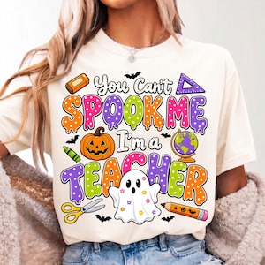May include: A cream-colored t-shirt featuring a colorful graphic with the text "You Can't SPOOK ME I'm a TEACHER". The design includes a pumpkin, a globe, scissors, a pencil, a ruler, an eraser, and a ghost decorated with small colorful flowers.
