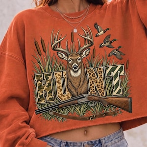 May include: An orange long-sleeve sweatshirt featuring a graphic print of a deer head, cattails, and three flying ducks. The word HUNT is printed in large block letters with camouflage and leopard patterns. A shotgun is positioned below the deer.