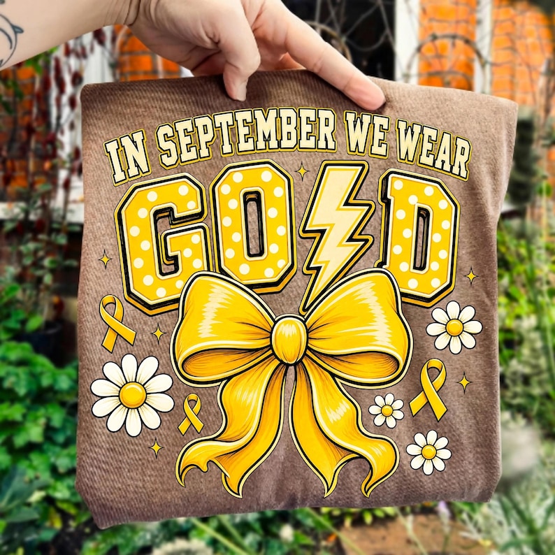 May include: A brown t-shirt featuring the text IN SEPTEMBER WE WEAR GOLD in yellow block letters with white polka dots. The letter L is replaced by a yellow lightning bolt. A large yellow bow, small yellow ribbons, and white daisies are printed.