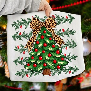 Op de afbeelding: Een wit sweatshirt met een afbeelding van een groene kerstboom versierd met rode kerstballen en bekroond met een grote strik met luipaardprint. Het ontwerp is omringd door horizontale rijen groene dennentakken en kleine rode lampjes.