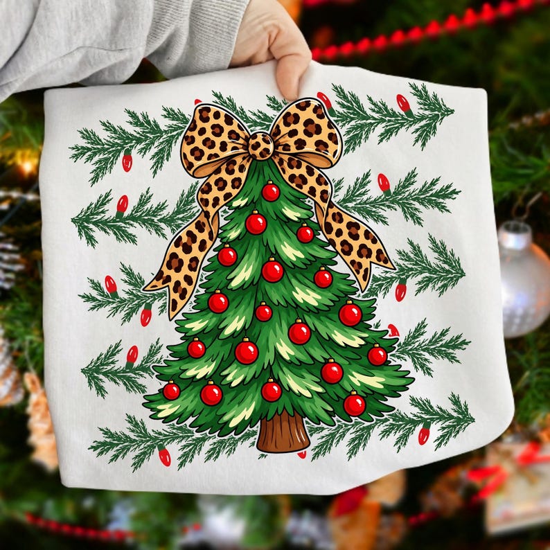 Op de afbeelding: Een wit sweatshirt met een afbeelding van een groene kerstboom versierd met rode kerstballen en bekroond met een grote strik met luipaardprint. Het ontwerp is omringd door horizontale rijen groene dennentakken en kleine rode lampjes.