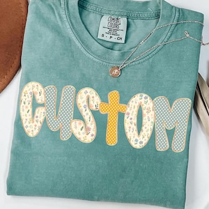 May include: A teal Comfort Colors t-shirt with the word "CUSTOM" in a floral and polka dot design. The letter "O" in "CUSTOM" is a yellow cross. A silver necklace with a pendant is on the shirt.