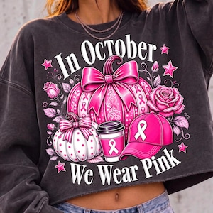 May include: A charcoal gray cropped sweatshirt featuring a graphic with the text In October We Wear Pink. The design includes two pumpkins, a coffee cup, a baseball cap, and roses, all in shades of pink with white breast cancer awareness ribbons.