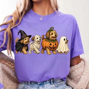 Puede incluir: Una camiseta de color morado claro con un gr&aacute;fico de cuatro perros peque&ntilde;os con disfraces de Halloween. Los perros est&aacute;n vestidos de bruja con sombrero y capa negra, momia con vendas blancas, calabaza y fantasma bajo una s&aacute;bana blanca.