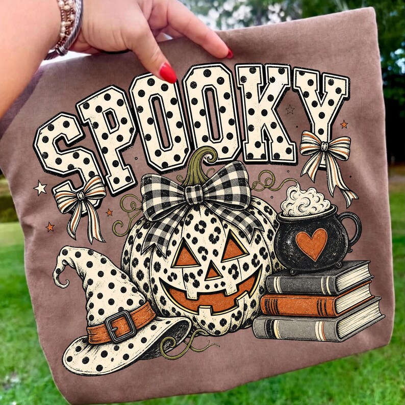 May include: A brown t-shirt featuring a graphic print with the word SPOOKY in white letters with black polka dots. Below, a pumpkin with a leopard print pattern, a black and white plaid bow, a witch hat, a cauldron with foam, and three books.
