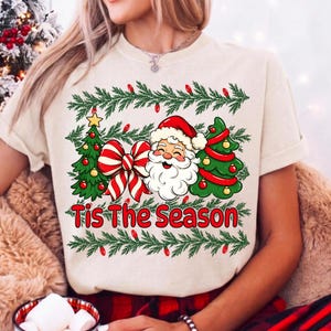 Puede incluir: Una camiseta de color crema con un estampado gr&aacute;fico de Santa Claus, dos &aacute;rboles de Navidad decorados y un gran lazo a rayas rojas y blancas. El texto 'Tis The Season' est&aacute; impreso en rojo debajo del gr&aacute;fico, enmarcado por guirnaldas de pino.