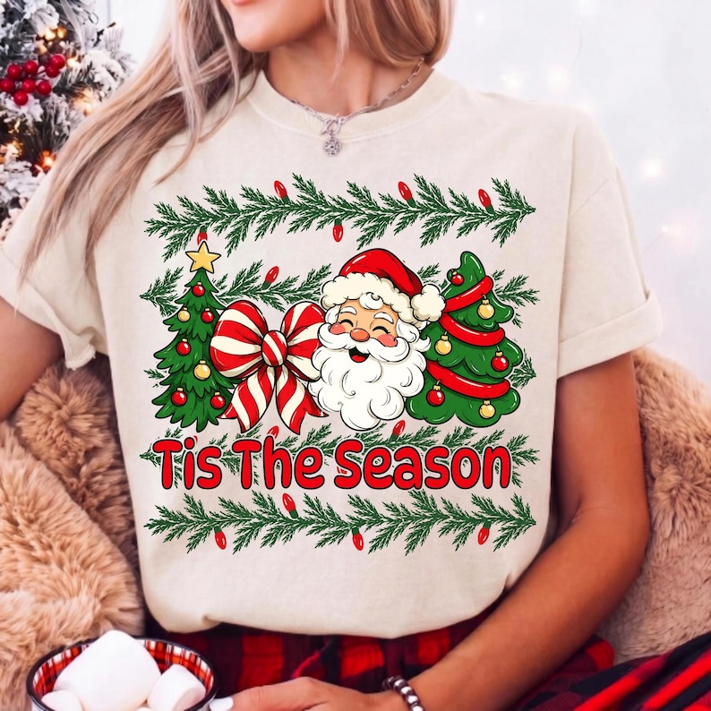 Puede incluir: Una camiseta de color crema con un estampado gr&aacute;fico de Santa Claus, dos &aacute;rboles de Navidad decorados y un gran lazo a rayas rojas y blancas. El texto 'Tis The Season' est&aacute; impreso en rojo debajo del gr&aacute;fico, enmarcado por guirnaldas de pino.