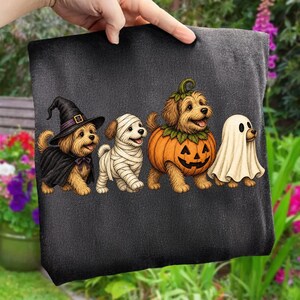Puede incluir: Una camiseta negra con un gr&aacute;fico de cuatro perros peque&ntilde;os con disfraces de Halloween. Los perros est&aacute;n vestidos de bruja con sombrero y capa negra, momia envuelta en vendas blancas, calabaza y fantasma cubierto con una s&aacute;bana blanca.