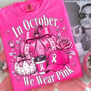 May include: A bright pink t-shirt featuring a graphic print with the text "In October We Wear Pink". The design includes two pumpkins, a rose, a coffee cup, and a baseball cap, all decorated with pink awareness ribbons and star accents.