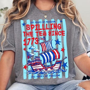 Puede incluir: Una camiseta gris jaspeado con un gr&aacute;fico de un barco con velas rojas, blancas y azules. El texto dice 250 YEARS OF FREEDOM, SPILLING THE TEA SINCE 1773 y 250 YEARS OF FREEDOM. El dise&ntilde;o incluye estrellas azules y rayas verticales azul claro.