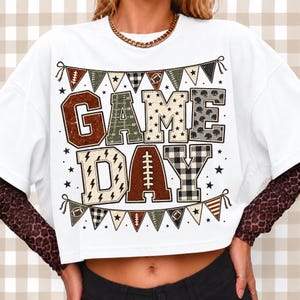May include: A white short-sleeved t-shirt featuring the text GAME DAY in large block letters with various patterns including footballs, stars, and plaid. The design is framed by triangular pennant banners and small stars on a white background.