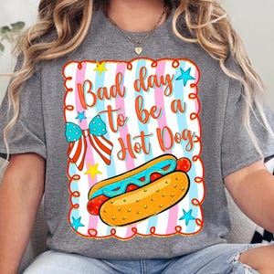 May include: A heather gray t-shirt featuring a graphic with the text "Bad day to be a Hot Dog" in orange script. The design includes a hot dog, a blue and white star-patterned bow, yellow stars, and a background of pink and blue vertical stripes.