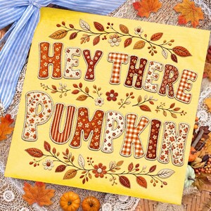 May include: A square yellow sign featuring the text HEY THERE PUMPKIN in large, block-style letters filled with various autumn-themed patterns including orange and brown florals, stripes, and gingham checks, surrounded by embroidered leaf motifs.