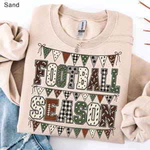 May include: A sand-colored crewneck sweatshirt featuring a graphic print of the text FOOTBALL SEASON in block letters. The design includes rows of triangular pennant banners with various patterns and football-themed icons in brown and green.