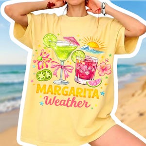 May include: Yellow t-shirt with a colorful graphic of two margarita cocktails, a sun, and ocean waves. The text "Margarita Weather" is printed below the drinks. The design includes lime slices, pink bows, and floral accents.