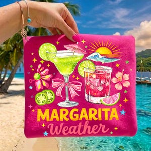 May include: A hot pink beach towel featuring a margarita and a cocktail with ice cubes. The towel has the words "Margarita Weather" in yellow and white. The background shows a beach scene with palm trees and the ocean.