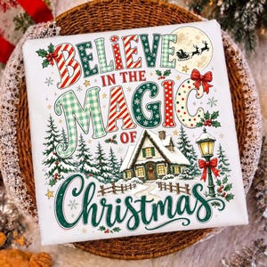 May include: A white t-shirt featuring the text "BELIEVE IN THE MAGIC OF CHRISTMAS" in red, green, and white patterns. The design includes a snowy cottage, pine trees, a glowing street lamp with a red bow, and a silhouette of Santa in a moon.