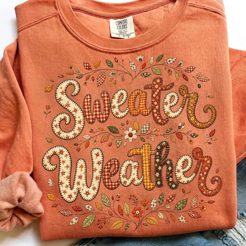 Pu&ograve; includere: Una felpa girocollo color corallo con la scritta Sweater Weather in un carattere decorativo a motivi vari. Le lettere sono riempite con stampe a tema autunnale, circondate da foglie, viticci e piccoli fiori illustrati.