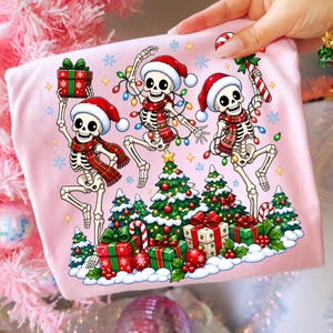 Op de afbeelding: Een lichtroze t-shirt met een afbeelding van drie skeletten met kerstmutsen en geruite sjaals, dansend rond kerstbomen en cadeaus. De skeletten zijn versierd met kleurrijke kerstverlichting en het ontwerp bevat sneeuwvlokken en sterren.
