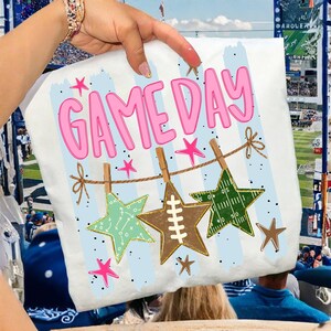 May include: A white t-shirt featuring the pink text GAME DAY above a graphic of a brown string with three hanging stars. The stars include a light green one with white play diagrams, a brown football-textured star, and a dark green football field star.