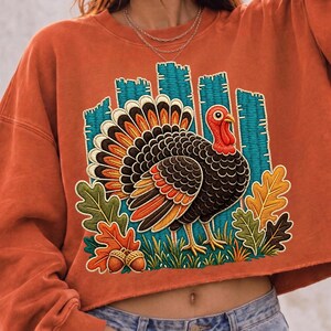 May include: A burnt orange cropped sweatshirt featuring a large embroidered graphic of a turkey with a fanned tail in shades of brown, orange, and yellow. The design includes teal vertical fence-like slats, green grass, and colorful autumn oak leaves.