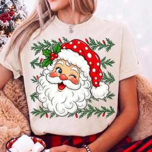 May include: A cream-coloured t-shirt featuring a graphic of a winking Santa Claus wearing a red polka-dot hat with a white pom-pom. The design includes green pine branches and small red lights surrounding the Santa face and holly leaves.