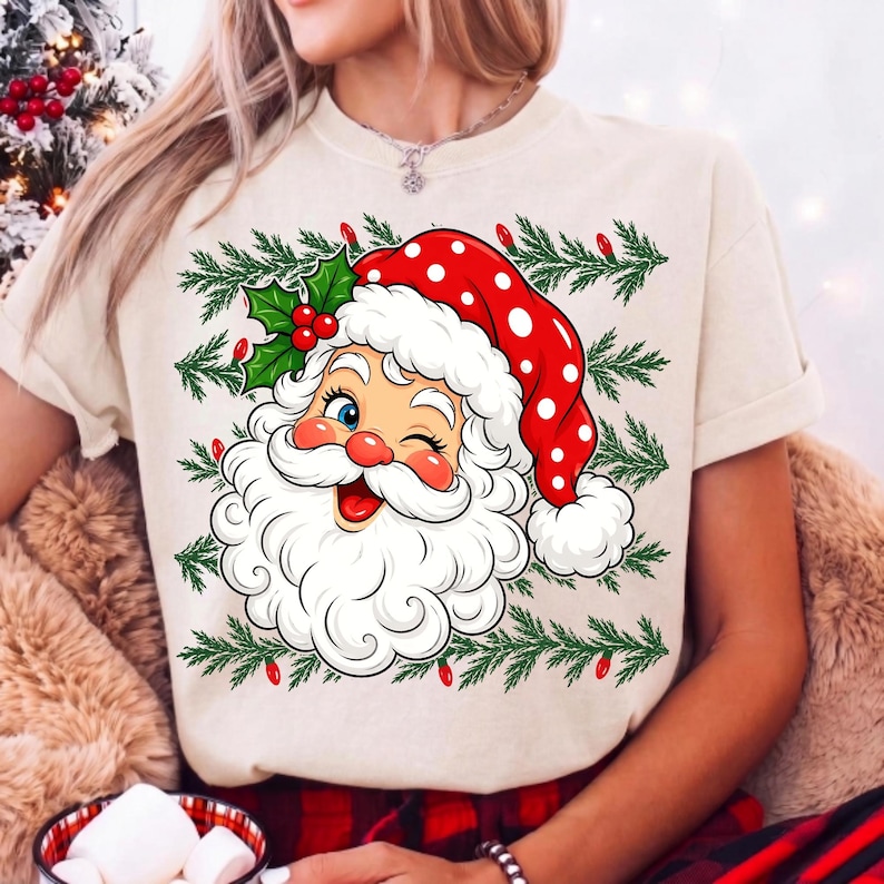 May include: A cream-coloured t-shirt featuring a graphic of a winking Santa Claus wearing a red polka-dot hat with a white pom-pom. The design includes green pine branches and small red lights surrounding the Santa face and holly leaves.
