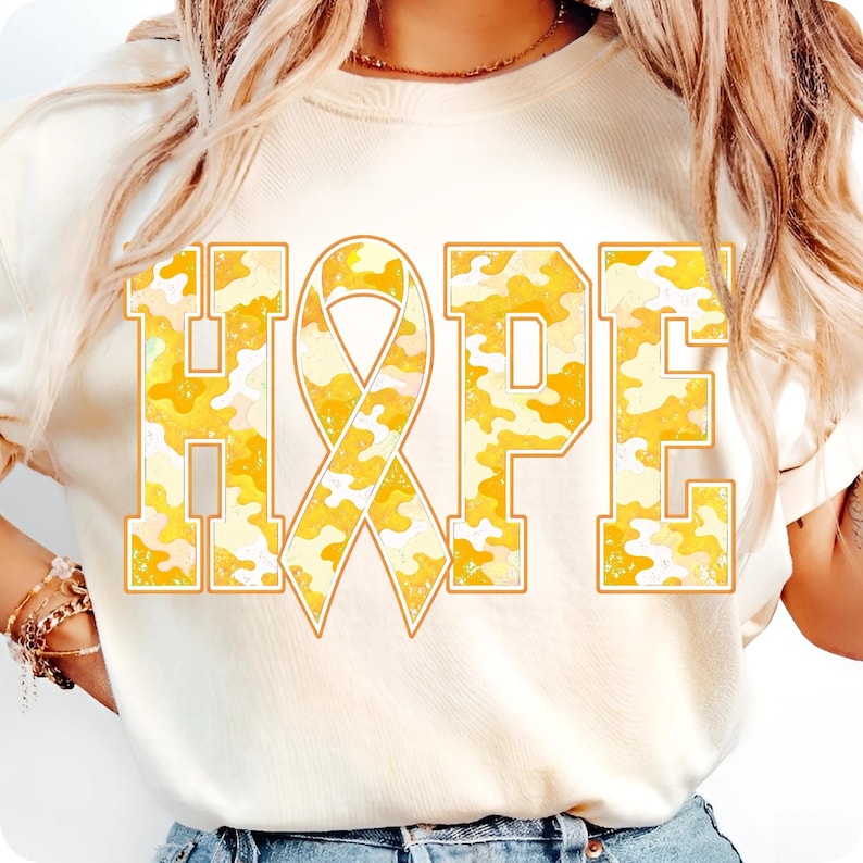 May include: A cream-coloured t-shirt featuring the word HOPE printed in large block letters. The letter O is replaced by a ribbon symbol. The text has a yellow and white camouflage pattern with a thin orange outline around each letter.
