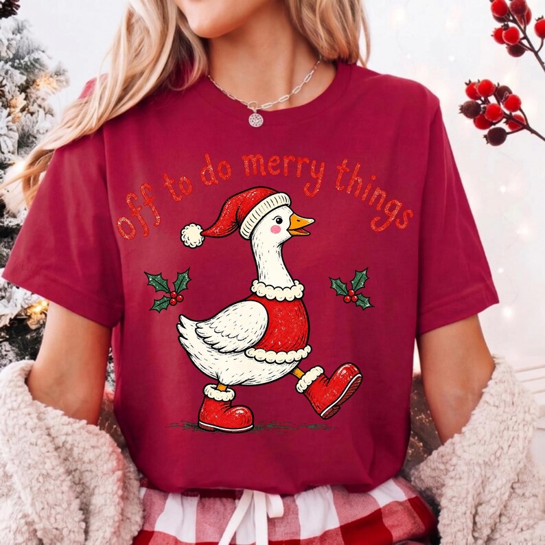 May include: A maroon short-sleeved t-shirt featuring a graphic of a white goose wearing a red Santa hat, a red vest, and red boots. The text "off to do merry things" is printed in orange above the goose, flanked by small green holly sprigs.