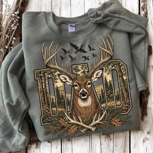 May include: A sage green crewneck sweatshirt featuring a graphic print of a deer head with large antlers centred over the word DAD. The letters are filled with a forest landscape scene, and the design includes flying ducks and pine branches.
