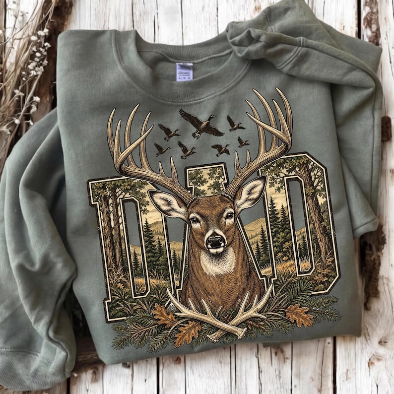May include: A sage green crewneck sweatshirt featuring a graphic print of a deer head with large antlers centred over the word DAD. The letters are filled with a forest landscape scene, and the design includes flying ducks and pine branches.