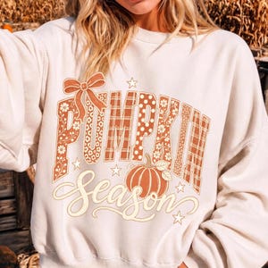 May include: A cream-colored crewneck sweatshirt featuring a graphic print with the text "PUMPKIN Season". The word "PUMPKIN" is in a block font with varied patterns including floral, polka dots, and plaid, topped with a bow. A pumpkin illustration sits below.