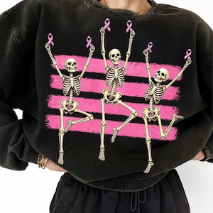May include: A black crewneck sweatshirt featuring a graphic print of three skeletons in dynamic poses. Each skeleton holds a small pink ribbon above its head. Behind the skeletons are four horizontal, distressed pink brushstroke stripes.