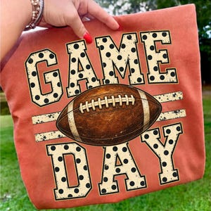 Op de afbeelding: Een roestoranje t-shirt met de tekst GAME DAY in grote, cr&egrave;mekleurige blokletters met zwarte stippen. Een bruine American football met witte veters en strepen is horizontaal geplaatst tussen de woorden GAME en DAY.