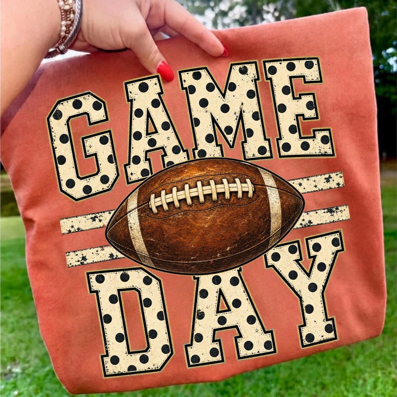 Op de afbeelding: Een roestoranje t-shirt met de tekst GAME DAY in grote, cr&egrave;mekleurige blokletters met zwarte stippen. Een bruine American football met witte veters en strepen is horizontaal geplaatst tussen de woorden GAME en DAY.