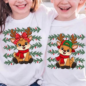 May include: Two white t-shirts featuring cartoon reindeer graphics. One reindeer wears a red polka-dot bow and red scarf, the other wears a green baseball cap and red scarf. Both are set against a background of green pine branches and red lights.