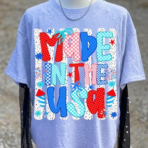 May include: A light grey t-shirt featuring a graphic print with the text MADE IN THE USA. The letters are filled with red, white, and blue patterns including polka dots and checkers, accented by firework illustrations and small blue bows.