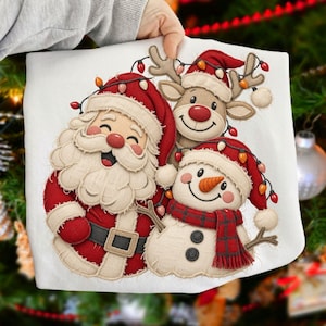 May include: A white t-shirt featuring a graphic of Santa Claus, a reindeer, and a snowman wearing red hats and scarves. The characters are decorated with a string of colorful Christmas lights. The design has a textured, layered appearance.