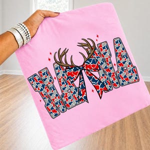 May include: A light pink fabric square featuring the word NANA in block letters filled with a red, white, and blue camouflage pattern. Above the letters are brown deer antlers and a red, white, and blue patterned bow with small red stars.