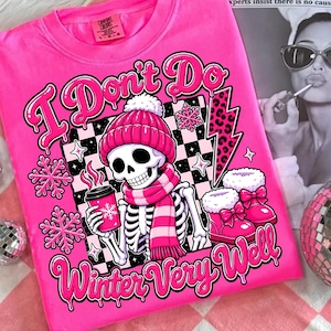 May include: A bright pink t-shirt featuring a graphic of a skeleton wearing a pink beanie and scarf, holding a pink coffee cup. The design includes pink snowflakes, a leopard print lightning bolt, and the text I Don't Do Winter Very Well.