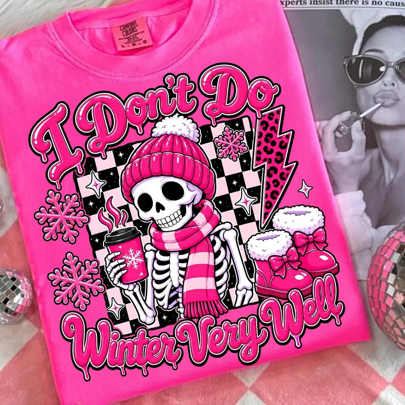 May include: A bright pink t-shirt featuring a graphic of a skeleton wearing a pink beanie and scarf, holding a pink coffee cup. The design includes pink snowflakes, a leopard print lightning bolt, and the text I Don't Do Winter Very Well.