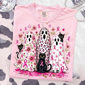 May include: A light pink t-shirt featuring a graphic of three ghosts and two black cats. The ghosts are decorated with pink floral, leopard print, and polka dot patterns. Pink breast cancer awareness ribbons and flowers surround the figures.