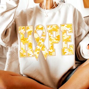 May include: A beige crewneck sweatshirt featuring the word HOPE printed in large block letters across the chest. The letter O is replaced by an awareness ribbon. The letters have a yellow and white camouflage pattern with a thin orange outline.