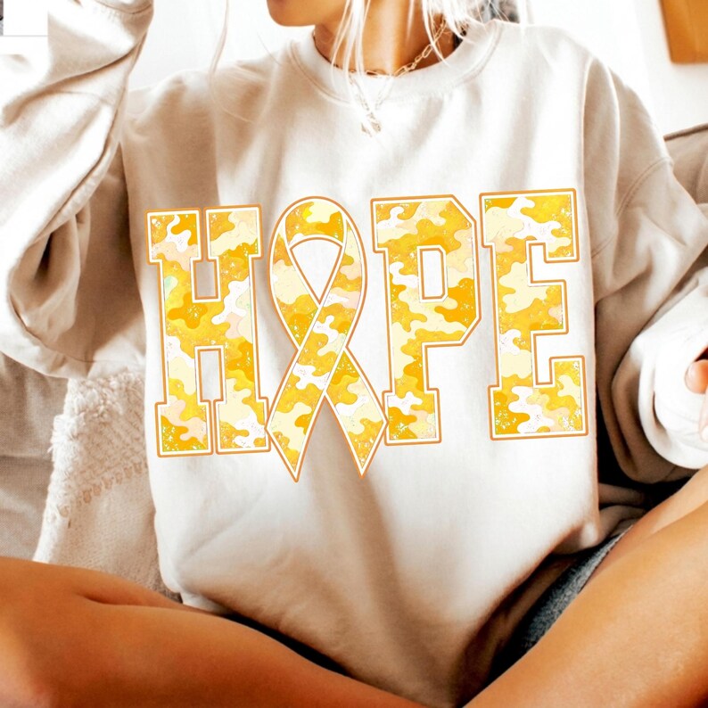 May include: A beige crewneck sweatshirt featuring the word HOPE printed in large block letters across the chest. The letter O is replaced by an awareness ribbon. The letters have a yellow and white camouflage pattern with a thin orange outline.